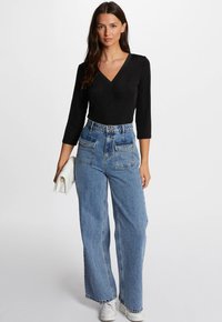 Schwarzer, langärmliger V-Ausschnitt-Bodysuit, kombiniert mit hochgeschnittenen, weiten, hellblauen Jeans. Das Model hält eine weiße Clutch. Lässiges Schuhwerk.