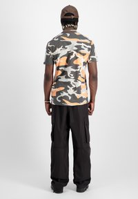 Man med pärlhår som bär en brun keps, en orange-grå camouflage t-shirt, svarta cargobyxor och svarta skor, stående med ryggen mot kameran.
