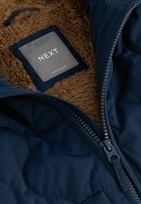 Marinblå quiltad jacka med en brun fuskpälskantning, framdragkedja och en grå etikett med texten "NEXT". Materialet verkar slätt och strukturerat.