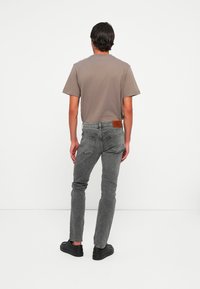 Man wearing gray slim-fit jeans, a brown short-sleeve t-shirt, and black shoes standing facing away against a plain white background.