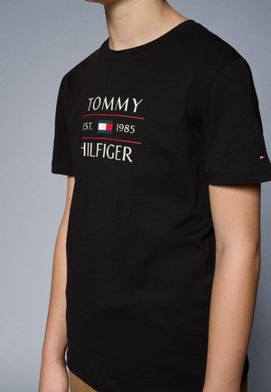 Person iført en sort Tommy Hilfiger t-shirt med logo og teksten "Est. 1985" foran samt en lille logo på ærmet.
