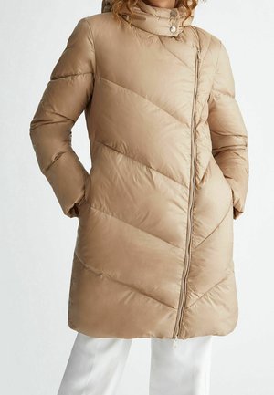 Manteau matelassé beige avec des coutures diagonales, col montant, fermeture éclair frontale, les mains dans les poches latérales, porté avec un pantalon blanc.