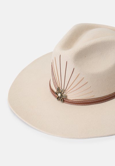 Breedgerande beige vilten hoed met een decoratieve bruine band. Heeft een metalen bloemenaccent en reliëflijnen die vanaf de kroon lopen.