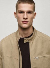 Beige suede jacket with a stand collar, side zip closure, and chest pocket. Worn over a dark knit sweater, featuring a smooth texture.