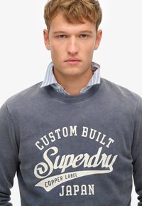 Sudadera de color azul marino con escote redondo y puños de canalé. Presenta un gran estampado gráfico en blanco que dice "CUSTOM BUILT Superdry JAPAN."