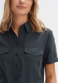 Dunkelgraues Kurzarm-Hemd mit umgeschlagenen Ärmeln, zwei Brusttaschen, glatter Textur und einem kragenförmigen Halsauschnitt. Verfügt über dezente Nähdetails.