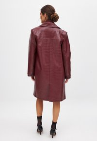 Cappotto in ecopelle color bordeaux con texture a coccodrillo, taglio oversize, ampio colletto, bottoni sui polsini e design lungo fino al ginocchio.