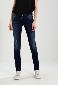 Jeans skinny blu con una texture sbiadita, cinque tasche e chiusura con bottone, abbinati a una maglietta nera e a delle sneakers bianche.