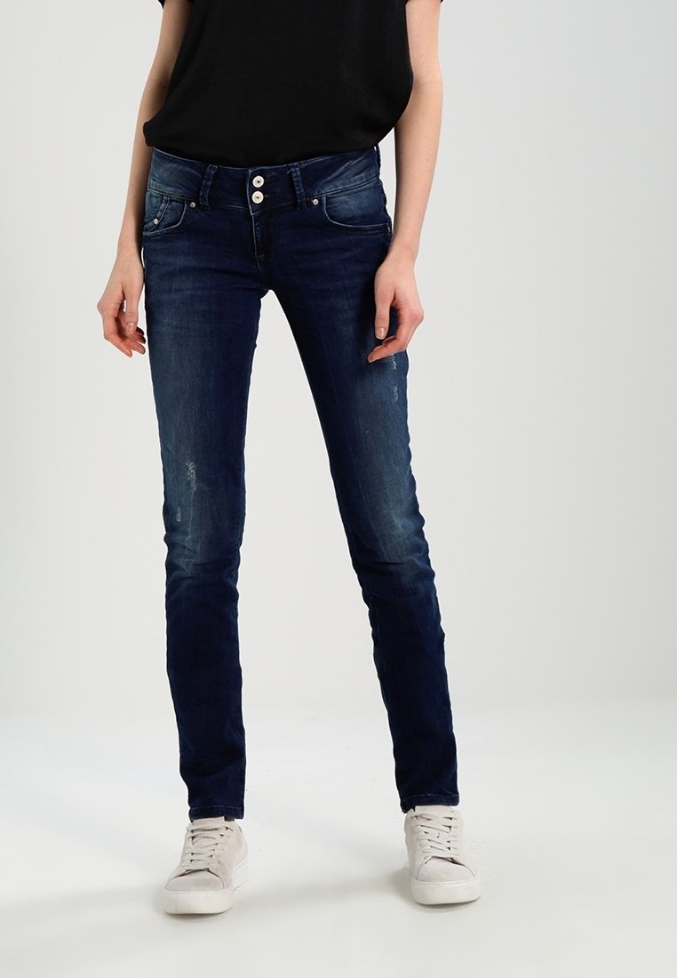 Jeans skinny blu con una texture sbiadita, cinque tasche e chiusura con bottone, abbinati a una maglietta nera e a delle sneakers bianche.