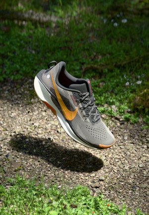 Šedá sportovní bota s texturovaným svrškem, oranžovým logem Swoosh a bílou mezipodešví, zavěšená nad kamenitou a travnatou plochou.