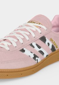Ροζ σουέτ αθλητικό παπούτσι Adidas με λευκά κορδόνια, ασπρόμαυρο ριγέ σχέδιο στο πλάι, σόλα από καουτσούκ και χρυσό λογότυπο "SPEZIAL" με καρδιά.