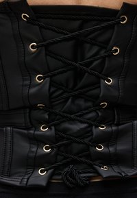 Μαύρο κορσέ με χρυσά οπές και μαύρα στριφογυριστά κορδόνια που διασταυρώνονται και δένονται σε κόμβο στο κάτω μέρος.