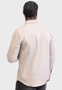 Jasnobeżowa koszula z długim rękawem, kołnierzykiem na guziki, detalem tyłu z karczkiem oraz prostym dołem; wykonana z teksturowanej bawełnianej tkaniny.