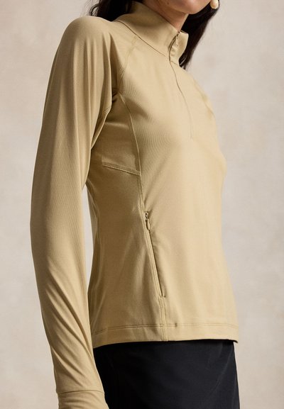 Μπεζ αθλητικό μακρυμάνικο τοπ με γιακά μισού φερμουάρ και τσέπη με φερμουάρ στο πλάι, που φοράει ένα άτομο που στέκεται μπροστά σε ουδέτερο φόντο.