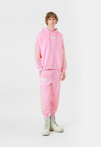 Sudadera rosa claro con detalles de alas bordados en las mangas, combinada con pantalones de chándal a juego que cuentan con un bolsillo lateral y un panel de acento.