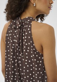 Femme aux cheveux bouclés portant une robe marron sans manches à pois blancs, nouée à l'arrière du cou, et des boucles d'oreilles argentées en forme de goutte.