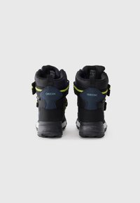 Zwarte mid-top sneakers met een textuur bovenwerk, voorzien van neon gele accenten en een solide zwarte zool. Merkenlogo zichtbaar op de hiel.