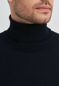 Pull à col montant côtelé bleu marine avec un col haut, texture tricotée et design ajusté, partiellement visible sur un fond neutre.