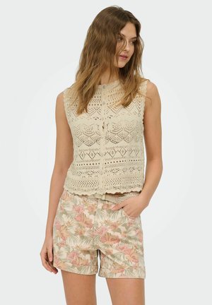 Nő laza hullámos hajjal, ujjatlan bézs horgolt felsőt és virágmintás rövidnadrágot visel, kezét a zsebében tartva, oldalra néz.