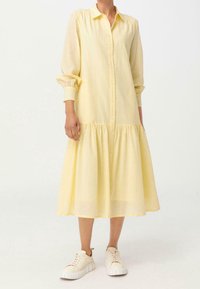 Żółta sukienka z długimi rękawami, z guzikami na całej długości, z zebranym, warstwowym dołem i lekkiego materiału. W połączeniu z białymi tenisówkami o swobodnym fasonie.