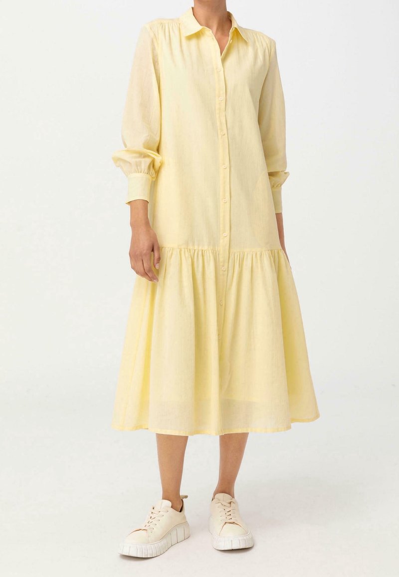 Żółta sukienka z długimi rękawami, z guzikami na całej długości, z zebranym, warstwowym dołem i lekkiego materiału. W połączeniu z białymi tenisówkami o swobodnym fasonie.