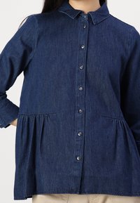 Denim shirt in dark blue with a button front, collar, and gathered detail at the waist. Features long sleeves with buttoned cuffs.