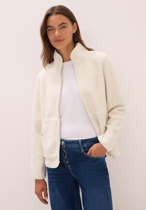 Frau in einer weißen Fleece-Zip-Jacke über einem weißen Shirt und blauen Jeans, stehend vor einem hellen, einfarbigen Hintergrund.