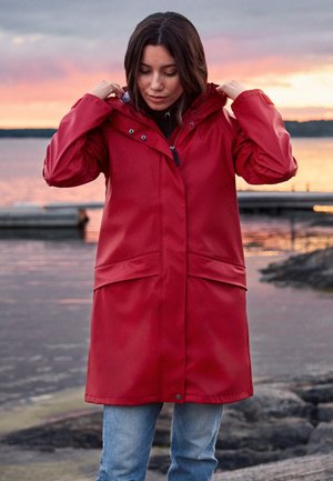 Kobieta poprawiająca kaptur długiego czerwonego płaszcza przeciwdeszczowego, stojąca przy wodzie o zachodzie słońca, w tle pomost i skaliste wybrzeże.