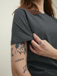 Camiseta de algodón gris oscuro de manga corta, con tatuajes visibles en el brazo, que incluyen varios diseños y texto.