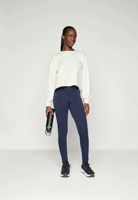 Ανοιχτό γκρίζο κοντό φούτερ, ναυτικές κολάν και μαύρα αθλητικά παπούτσια. Το μοντέλο κρατάει ένα μαύρο μπουκάλι νερού. Λείο ύφασμα, στενή εφαρμογή.