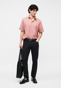 Νέος άνδρας που φοράει κοντομάνικο ροζ πουκάμισο με σχέδια, μαύρα τζιν, μαύρη ζώνη, μαύρα μοκασίνια, κρατώντας μια μεγάλη μαύρη τσάντα ώμου.