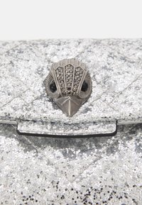 Stříbrná třpytivá kabelka s prošívaným designem. Vybavena kovovým zapínáním ve tvaru ptáka s klenotnickými akcenty a černými detaily na očích.