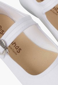Zapatos de tela blanca con punta redonda, tiras cruzadas con una hebilla metálica y una plantilla texturizada de color marrón. Superficie lisa y suela plana.