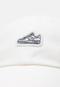 Weißes Baseballcap mit einer gestickten schwarzen Kontur eines Sneakers auf der Vorderseite. Hergestellt aus einem Stoff mit glatter Textur.