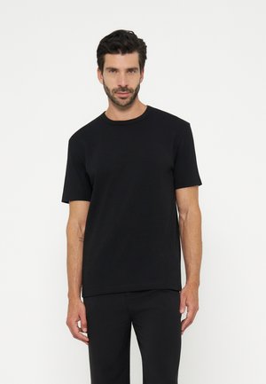 Homme aux cheveux foncés et à la barbe, portant un t-shirt noir uni à manches courtes et un pantalon noir, debout devant un fond blanc.