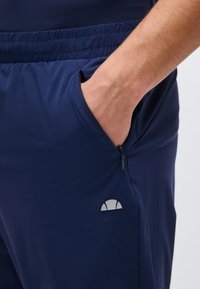 Χέρι που μπαίνει στην τσέπη με φερμουάρ σε σορτς με ελαστική μέση σε ναυτικό μπλε χρώμα και μικρό λευκό λογότυπο στο μηρό.