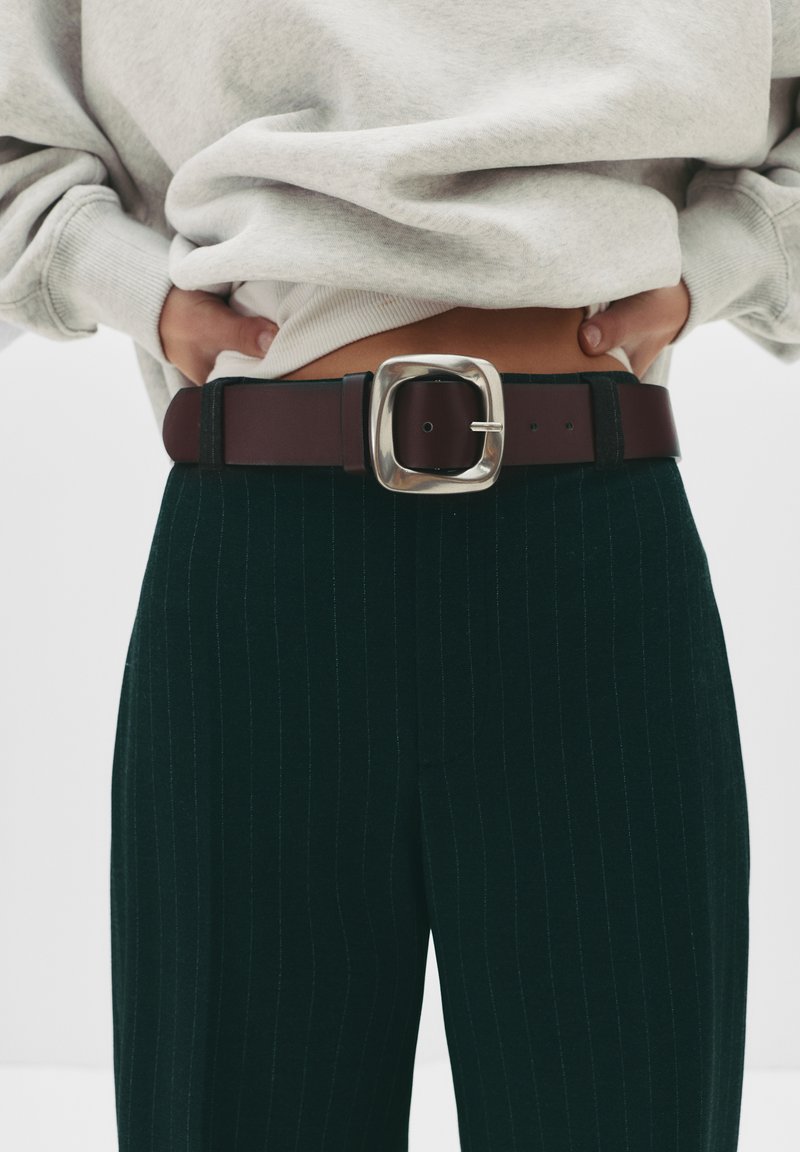 Barna bőröv fényes, négyzet alakú ezüstcsattal, zöld csíkos nadrágon viselve, részben egy szürke pulóver takarja.