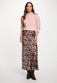 Vrouw in een roze trui met schouderknopen, een middenlange rok met luipaardprint en zwarte enkellaarsjes met hakken, staand tegen een witte achtergrond.