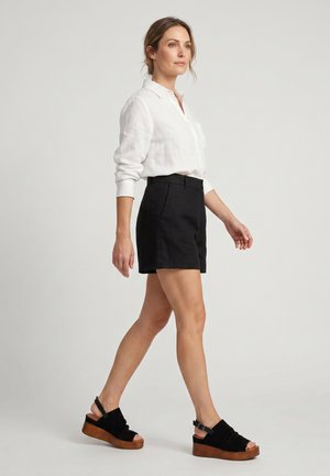 Frau läuft, trägt ein weißes Langarmshirt, schwarze Shorts mit hoher Taille und schwarze Keilsandalen mit Holzsohle vor einfachem Hintergrund.