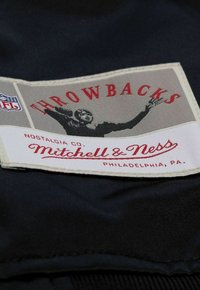 Das Mitchell & Ness Etikett zeigt ein schwarz-weißes Bild eines Spielers mit rotem Text, der "THROWBACKS" auf einem cremefarbenen Hintergrund liest, der an dunklem Stoff befestigt ist.