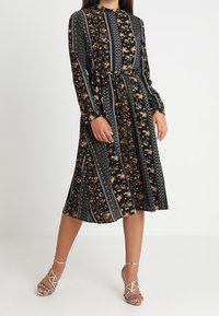 Robe midi noire à fleurs avec des manches longues, taille cintrée et motifs rayés. Présente des accents floraux orange et blancs. Portée avec des talons à lanières.