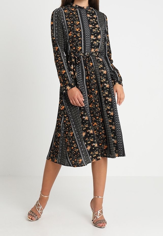 Robe midi noire à fleurs avec des manches longues, taille cintrée et motifs rayés. Présente des accents floraux orange et blancs. Portée avec des talons à lanières.