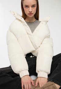 Biała kurtka puchowa z krzyżowym wzorem, z zamkiem z przodu i elastycznymi mankietami, noszona na szarej koszuli, o matowym wykończeniu i puszystej teksturze.