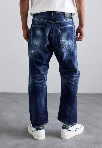 Pleekinud tume sinine denim teksapükk, millel on lõdvestatud istuvus, kahe tagatasku ja kulumise üksikasjad. Kandest valged tossud.