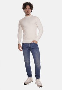 Maglione aderente a maniche lunghe di materiale morbido, abbinato a jeans blu e sneakers bianche. Il modello sta in piedi dritto con le mani rilassate.