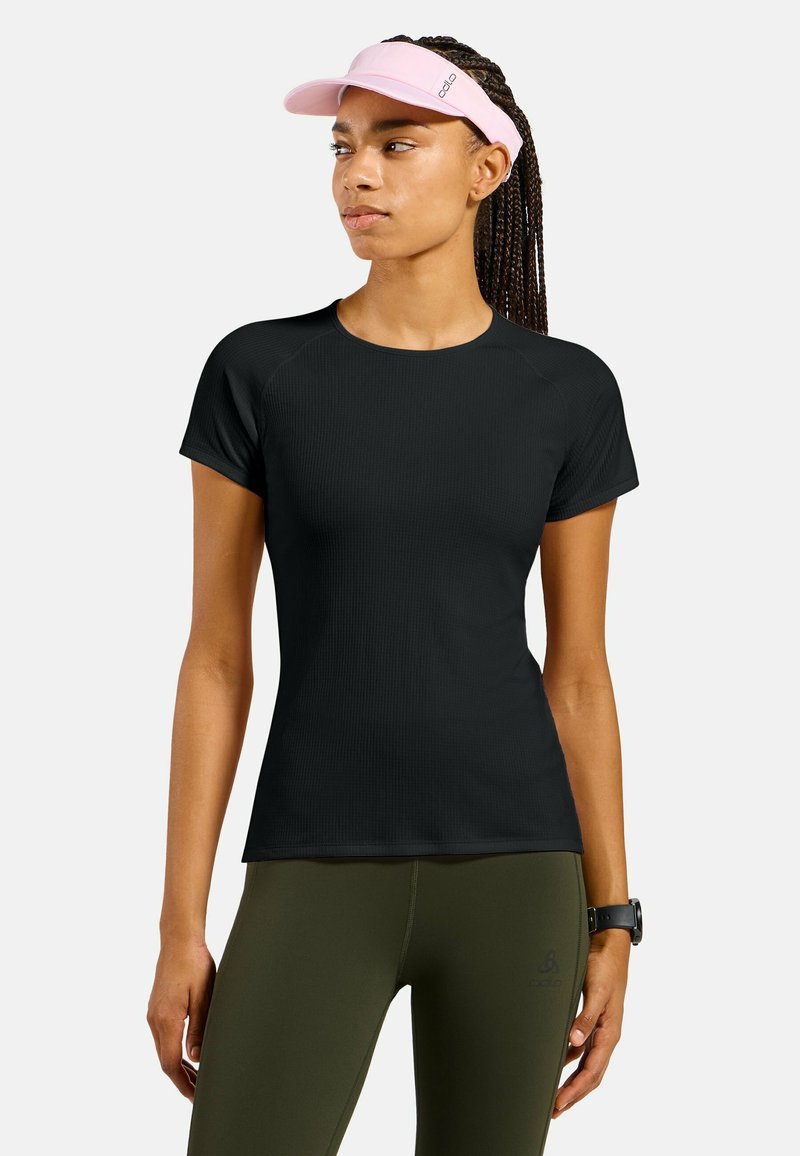 Femme aux cheveux tressés portant un t-shirt de sport noir à manches courtes, des leggings vert olive, une visière rose et une montre de fitness noire.