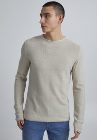 Helles beiges Langarmshirt mit strukturiertem Strickdesign und Rundhalsausschnitt, kombiniert mit blauen Jeans.