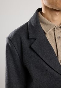 Cappotto di lana grigio scuro con colletto a revers, indossato su una camicia beige con bottoni in feltro. Il tessuto ha una consistenza liscia e un design classico.