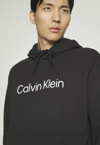 Black hoodie made of soft fabric, featuring a white "Calvin Klein" logo across the chest, adjustable drawstring hood, and front pocket.