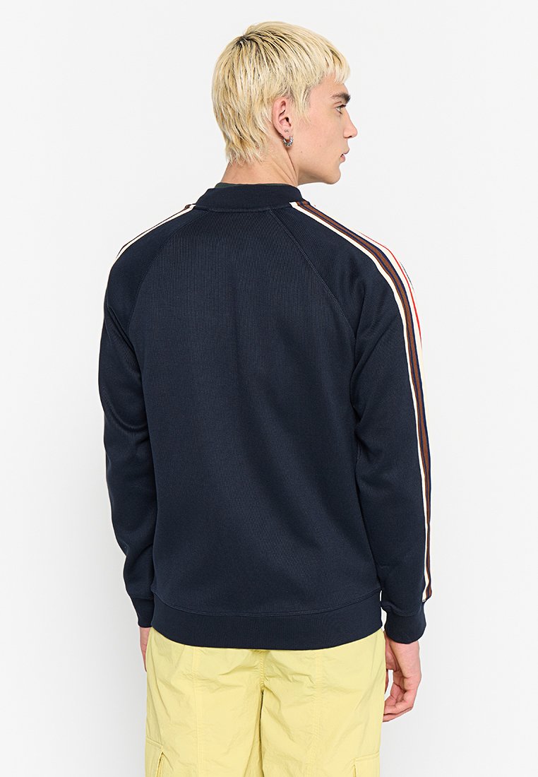 Eine blonde Person steht mit dem Rücken zur Kamera, trägt einen navyblauen Sweatshirt mit gestreiften Ärmeln und hellgelben Hosen.